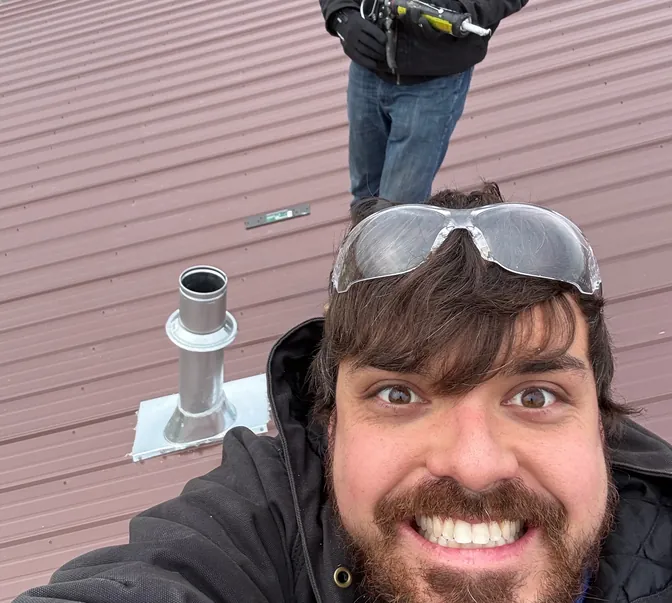 Cheerful BreezeTech Heating and Cooling technician on a metal roof beside a new furnace vent