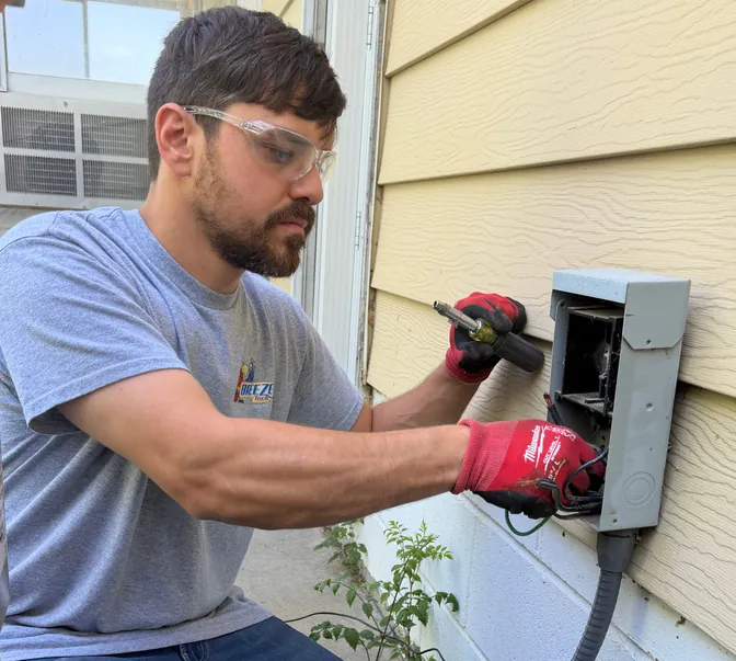 A BreezeTech technician in safety glasses wiring an AC disconnect box on the side of a home