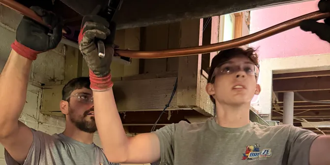 Two BreezeTech Heating and Cooling technicians installing refrigerant lines above a furnace