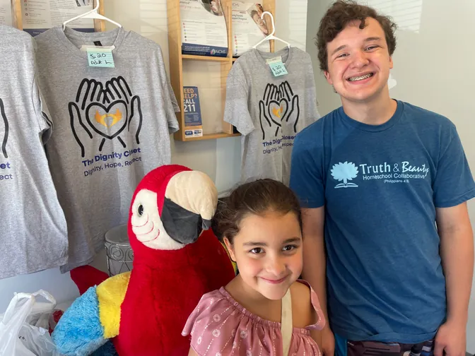 Two kids with Max the Macaw beside The Dignity Closet T-shirts