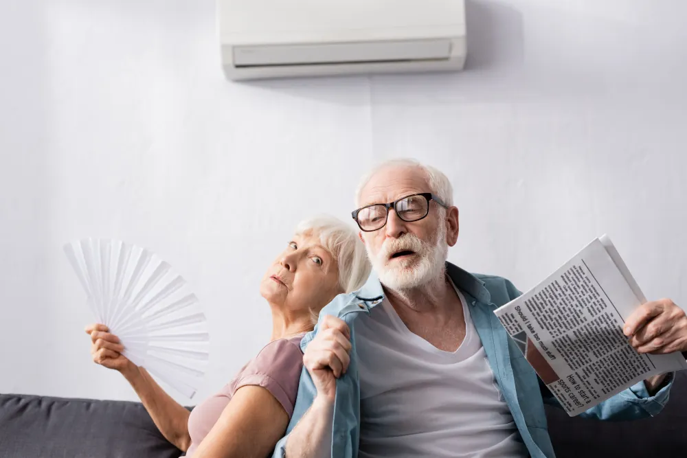 Senior man holding newspaper near tired wife with fan under ductless AC in living room